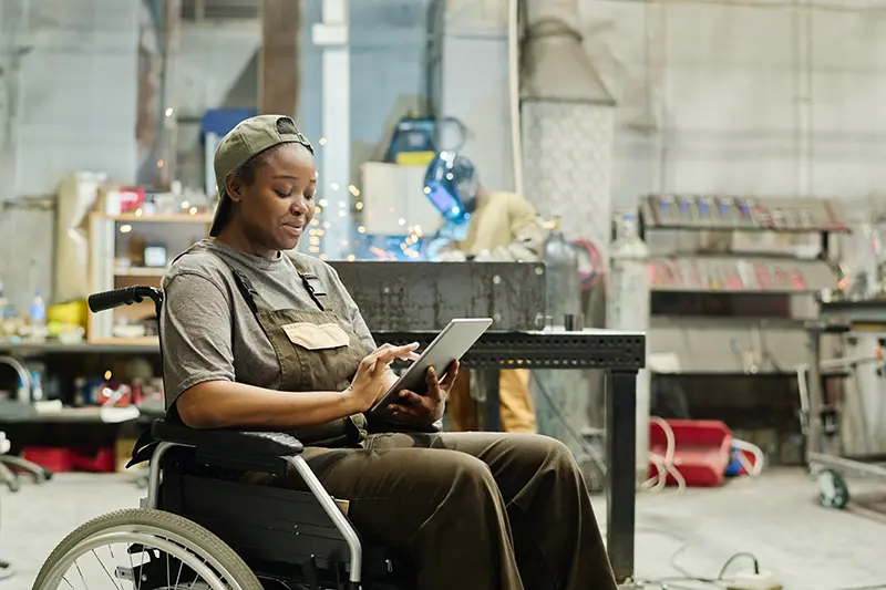 Frau mit dunkler Hautfarbe und Käppi in einer Werkstatt. Sitzt im Rollstuhl und hält ein Tablet in der Hand